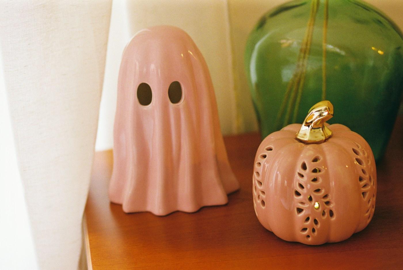 A pink ceramic ghost beside a pink pumpkin lantern with a gold stem, with a green vase in the background